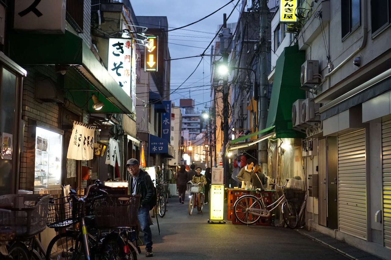 our-story A bustling evening street scene in a narrow Japanese alleyway with people and bicycles.