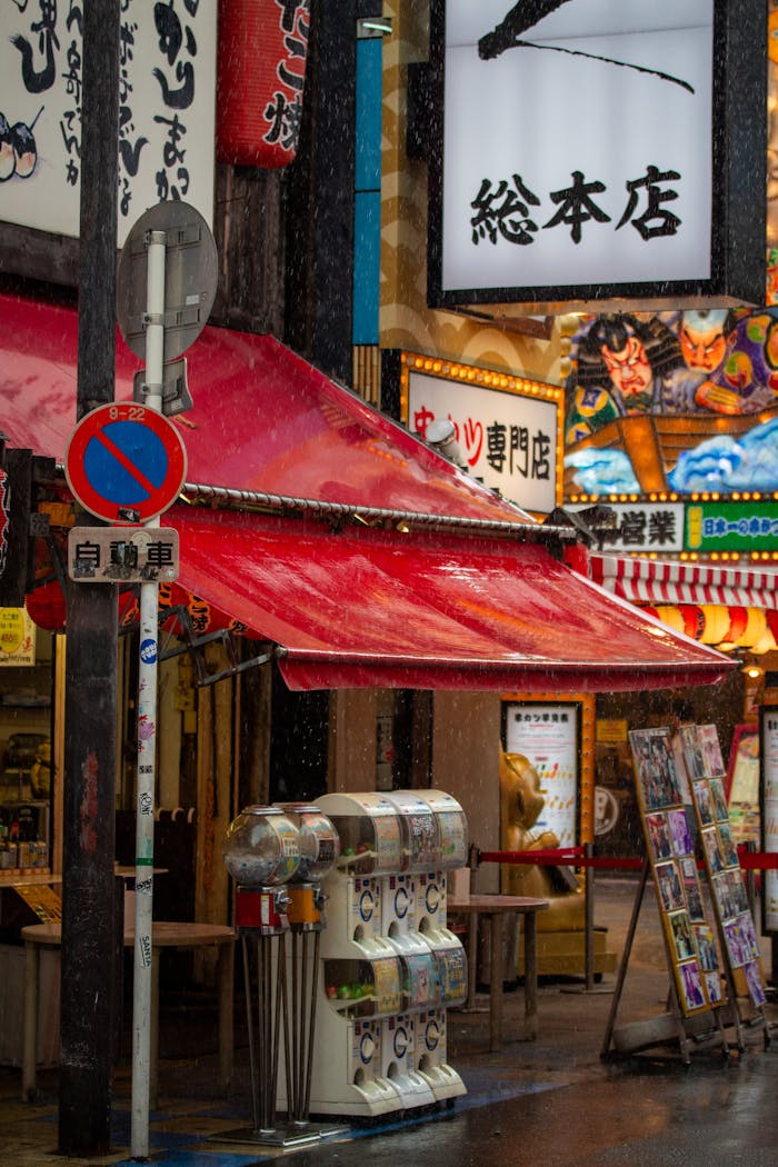 Vibrant city street scene with colorful shop signs and a red awning under rain.